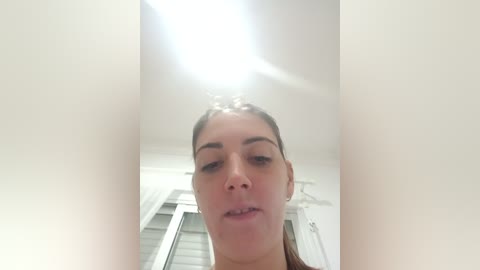 Video of a young woman with light skin and brown hair, standing in a bright, white room with a ceiling light shining overhead.