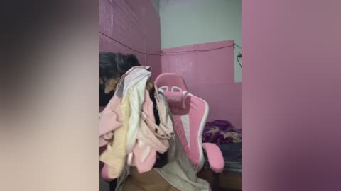 Video of a small, pink, child-sized gaming chair in a cluttered, dimly lit room with pink walls and a beige ceiling. The chair is surrounded by various clothes and objects.
