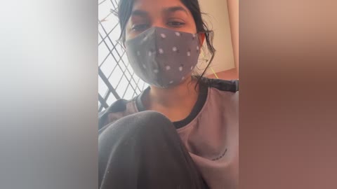Video of a young woman with medium skin tone, wearing a gray face mask with white polka dots, a gray sweatshirt, and gray pants. She is indoors near a window with a grid pattern.