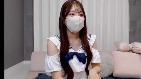 Video of an East Asian woman with long brown hair, wearing a white sailor outfit with a blue bow, mask, and pastel backdrop.