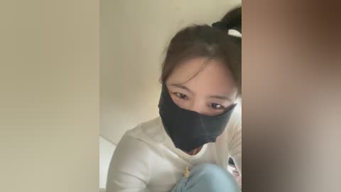 A video of a young Asian woman with dark hair tied in a ponytail, wearing a black mask and white top, crouched against a beige wall.
