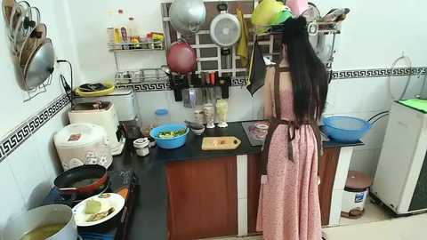 Media: Video of a woman with long black hair in a pink dress stirring a pot in a small, cluttered kitchen with white tiles and a black counter.