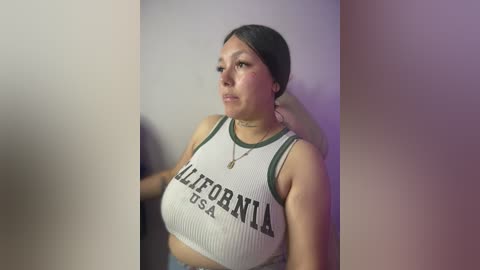 Video of a plus-sized Latina woman with a medium skin tone, wearing a white ribbed crop top that reads \"California USA\" in bold letters, and light blue jeans. She has dark hair tied back, and her expression is contemplative.