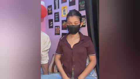 A video of an Asian woman with a mask, wearing a burgundy blouse, seated in a room adorned with colorful posters and a red balloon.