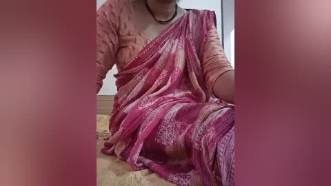 A video of a woman in a pink saree with intricate patterns, sitting on a wooden floor. She has a black beaded necklace and her dark hair is partially visible. The background is blurred, with a light wall.