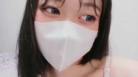 Video of an East Asian woman with straight black hair, wearing a white surgical mask, and a white spaghetti-strap top, against a plain white background.