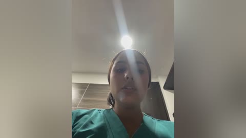 Video of a young woman with light brown skin, dark hair pulled back, wearing teal scrubs, taken from a low angle in a dimly lit room with a bright light source above her head.