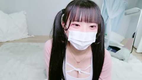 Video of a young Asian woman with long black hair in pigtails, wearing a white surgical mask and a pink cardigan over a white lace top, kneeling on a plush carpet in a bright, minimalistic room with white walls and a mirror.