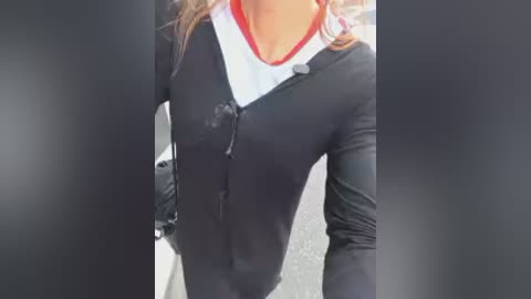 Video of a woman's torso wearing a black long-sleeve top with a white collar and red trim, hands in pockets, standing outdoors on a gray pavement.