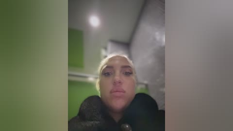 Video of a blonde woman with a neutral expression, wearing a black fur coat, indoors with a green wall and industrial ceiling lights in the background.