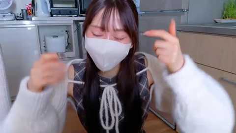 Video of an Asian woman with long dark hair and fair skin, wearing a white face mask and black plaid shirt, with white gloves, in a modern kitchen, gesturing with both hands.