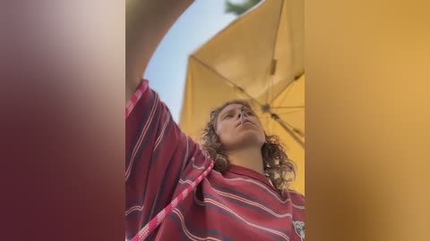 Video of a young woman with curly hair, wearing a red and white striped sweater, under a yellow umbrella. She gazes upwards, capturing an elevated perspective.