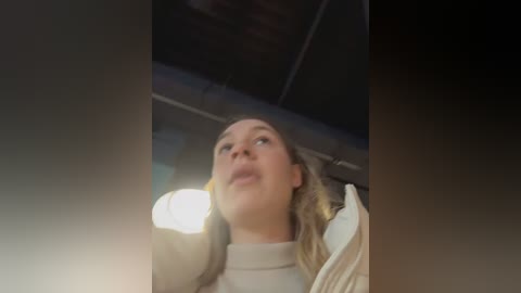 Video of a blonde woman with fair skin, wearing a beige sweater, looking upwards, possibly in a bathroom, with a blurry, dimly lit background.