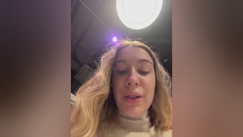 Video of a young woman with wavy blonde hair, wearing a cream-colored turtleneck sweater, standing in a dimly lit room with a round light fixture above. Her eyes are closed, and she has a relaxed expression.