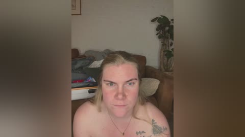 Video of a topless, pale-skinned woman with shoulder-length blonde hair, sporting a tattoo on her left shoulder, sitting on a couch in a cluttered living room.
