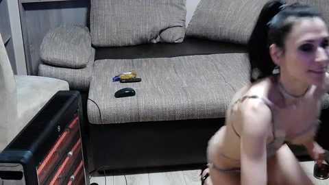 Video of a woman in lingerie kneeling on a wooden floor, with a beige sofa and electronics in the background, captured in a dimly lit room.