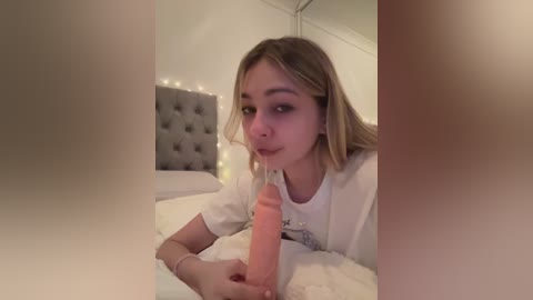 Video of a young woman with light skin and blonde hair, lying on a bed, holding a realistic, erect, pink dildo near her mouth. Background features a tufted gray headboard and string lights.