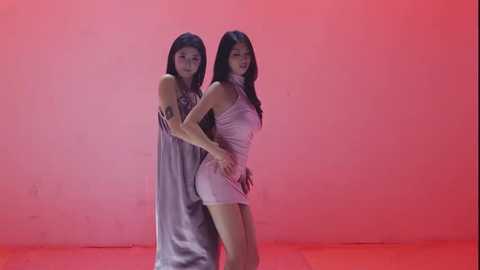 Video of two Asian women with long black hair, one in a silver dress, the other in a tight white dress, standing back-to-back against a pink wall, creating a striking, minimalist composition.
