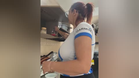 Video of a middle-aged woman with light skin, red hair tied back, wearing a tight white sports shirt, washing dishes in a kitchen with stainless steel appliances and cabinets.