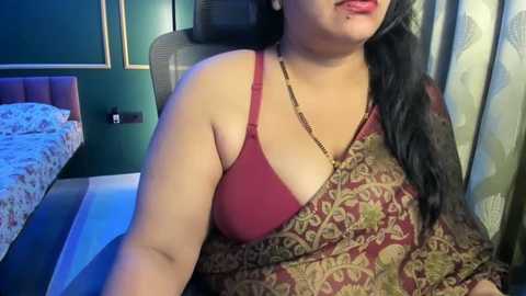 Video of a plus-size woman with medium skin tone, wearing a red lace bra and a paisley-patterned sari, seated on a bed in a dimly-lit room with teal walls and floral curtains.