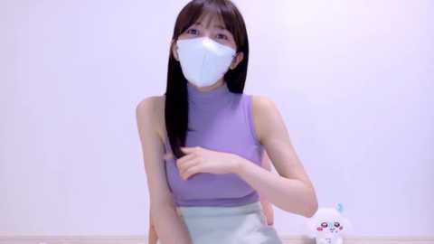 Video of an East Asian woman with long black hair, wearing a white mask, light purple sleeveless top, and light blue skirt, adjusting her hair. Background is a plain white wall with a white, cartoon character figurine.
