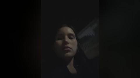 A dimly lit video captures a close-up of a woman's face with long dark hair, her eyes partially closed, and lips slightly parted, set against a dark background.