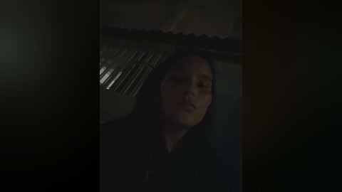 A dimly lit video shows a young woman with straight, dark hair lying on a bed with a metal headboard. Her face is partially illuminated, and she has a serene expression. The background is shadowy, with a hint of a window and blinds.