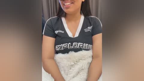 Video of a smiling woman with medium skin tone, medium-length black hair, wearing a dark gray athletic shirt with white accents and \"EXPLOSION\" text, and a white furry blanket. Background is a blurred, gradient brown to gray.