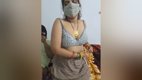 Video of a woman with medium skin tone, wearing a gray face mask, blue floral-patterned bra, and gray skirt, holding a yellow scarf, in a simple indoor setting.