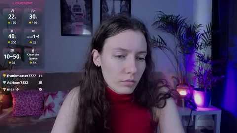Video of a young woman with long brown hair, wearing a red turtleneck, in a dimly lit room with a potted plant and framed pictures on the wall.