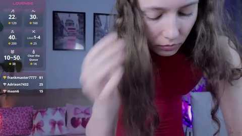 Video of a young woman with long brown hair, wearing a red sleeveless top, bending over, in a room with purple lighting and framed art on the wall.