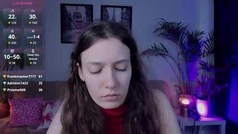 Video of a young woman with long brown hair, wearing a red turtleneck, in a dimly lit living room with plants, framed pictures, and a TV screen displaying her social media stats.