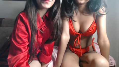 A video of two young women with long hair, one in a red satin robe, the other in a matching red bikini, sitting on a bed with a beige blanket.