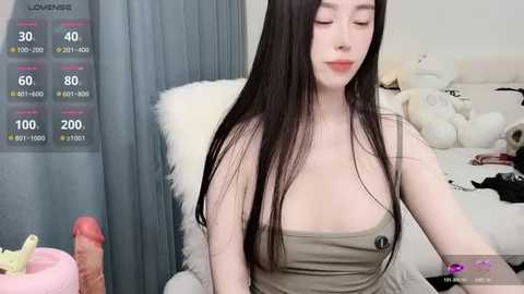 Video of a young Asian woman with long black hair, fair skin, wearing a strapless olive dress, sitting on a bed with plush toys.
