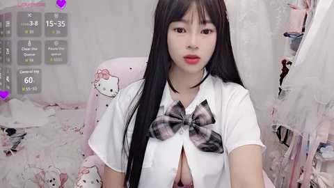 Video of a young Asian woman with long black hair, wearing a white schoolgirl outfit with a plaid bow. She sits in a pink Hello Kitty-themed room with a cluttered background.