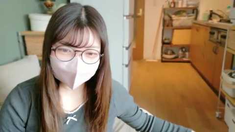 Video of a young woman with long brown hair, wearing glasses and a white face mask, standing in a modern kitchen with light wood cabinets and a white sink.
