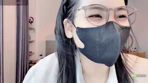 Video of an East Asian woman with long black hair, wearing clear glasses and a black face mask, looking into the camera in a well-lit room with neutral colors.