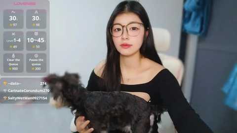 Video of a young Asian woman with long black hair, wearing glasses, black off-shoulder top, and holding a black curly dog. Background features a light-colored room with a blue garment hanging.