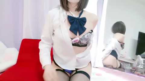 Video of an East Asian woman with a slender build, short black hair, wearing a white blazer, revealing a black lace bra and panties, garter belt, and thigh-high stockings. She sits on a red couch, in a room with white curtains and a mirror reflecting her.