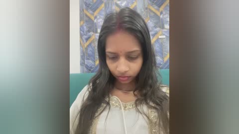 Video of a young South Asian woman with long black hair, wearing a white embroidered sari, sitting in a teal chair against a blue patterned wall. She appears contemplative.