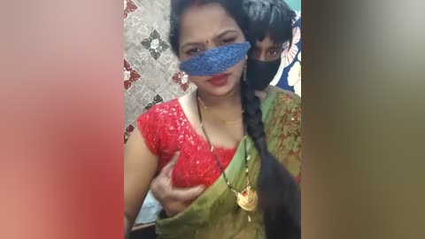 Video of a woman with medium-dark skin and black hair in a red sequined blouse, wearing a green saree. Her face is covered with a blue mask. Behind her, another person's face is partially visible.
