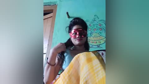 Video of a young woman with long black hair, wearing red sunglasses and a yellow and white checkered shawl, standing in a room with a teal-painted wall featuring a cartoonish drawing of a flying saucer.