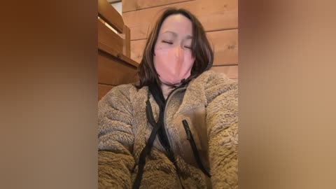Video of a woman with long brown hair, wearing a beige fleece jacket with black zippers, lying face down on wooden floor, her mouth covered with a pink mask, in a rustic wooden room.