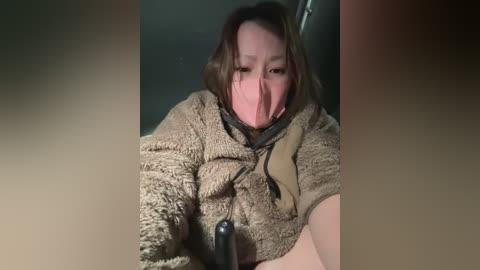 Video of a distressed woman with shoulder-length brown hair, wearing a beige fleece jacket and black mask, seated in a dimly lit room.