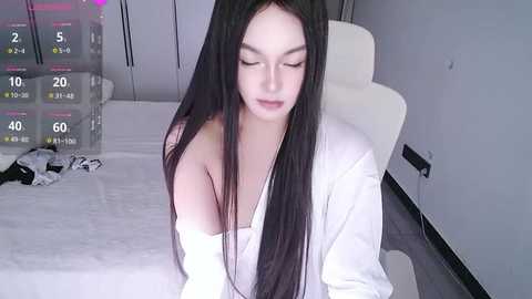 Video of a young woman with long black hair, pale skin, and closed eyes, wearing a white robe, sitting on a bed in a modern, minimalistic bedroom with white walls and a white chair.