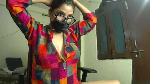 Media: A video of a woman in a colorful plaid shirt, black mask, and glasses, adjusting her hair in a modest, sparsely furnished room with wooden cabinets and a desk.