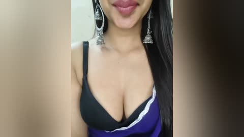 Video of a woman with medium skin tone, wearing a low-cut black and purple dress, revealing ample cleavage. She has long, straight black hair and wears large silver hoop earrings.