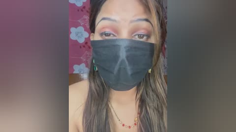 Video of a young woman with medium skin tone, wearing a black face mask, red lipstick, and a colorful beaded necklace, against a pink floral-patterned background.