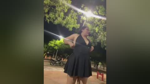 A video of a plus-size woman in a black halter dress, dancing under a bright, starry night sky, with trees and a bicycle in the background.