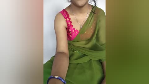 A video of a young woman with medium brown skin wearing a green sari with a pink, lacy blouse. She has a delicate gold necklace and a blue bangle on her wrist. The background is a gradient of white, green, and brown.